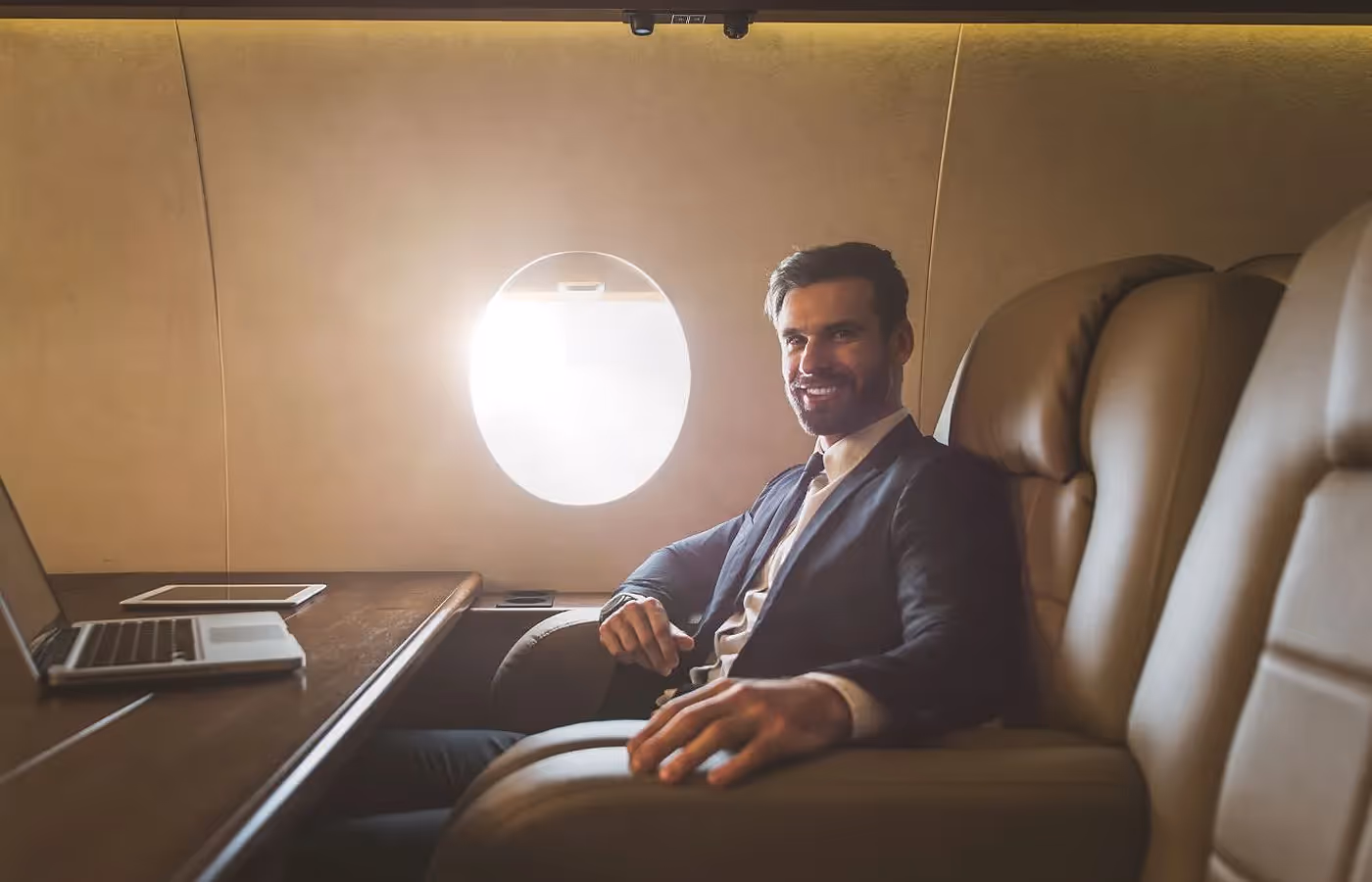 Smiling businessman in a suit sitting in a leather seat next to a window inside a private jet.