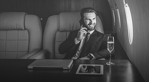Smiling man in a suit talking on a phone while sitting in a private jet with a glass of champagne on the table.