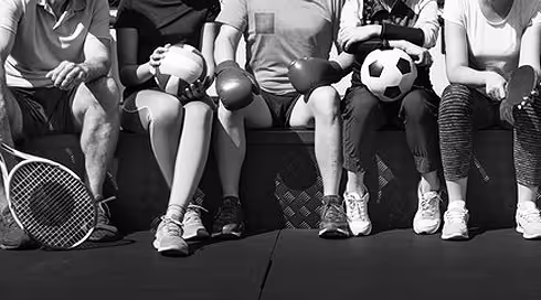 Five people sitting side by side holding different sports balls including a tennis ball, basketball, rugby ball, soccer ball, and volleyball, with a tennis racket on the ground.