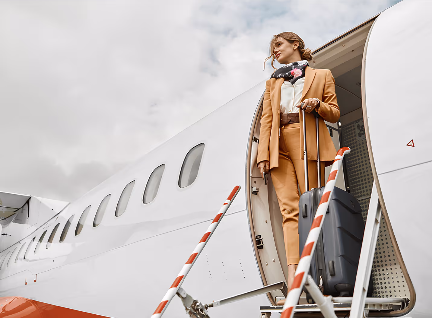 Woman in a tan suit with a scarf holding a suitcase standing at the entrance of a private jet.