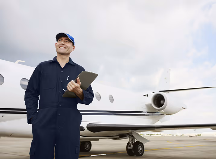 Smiling male aircraft mechanic in navy coveralls and cap holding a clipboard beside a private jet on the tarmac.