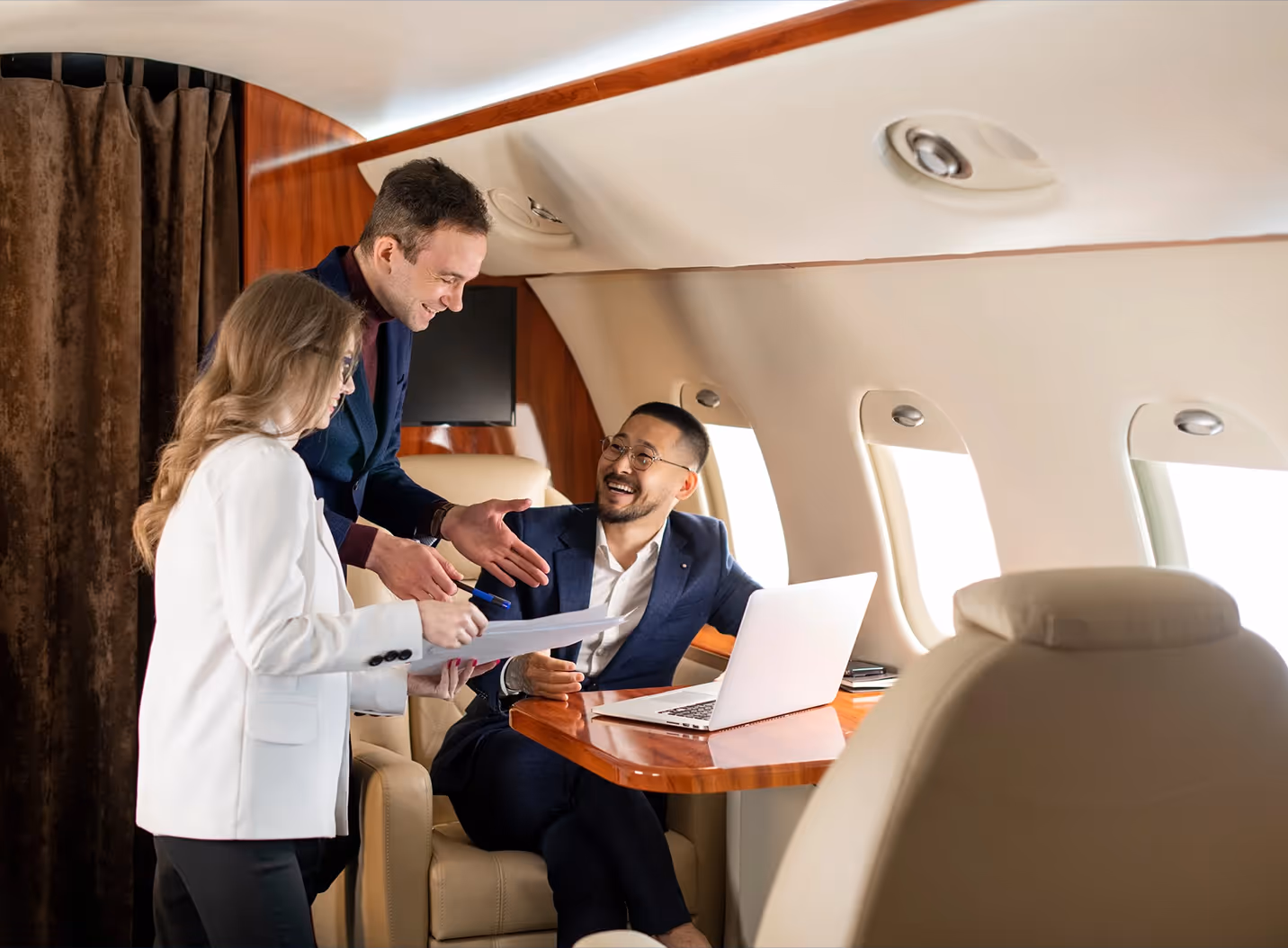 Three business professionals engaged in discussion inside a private jet with a laptop and documents on a wooden table.