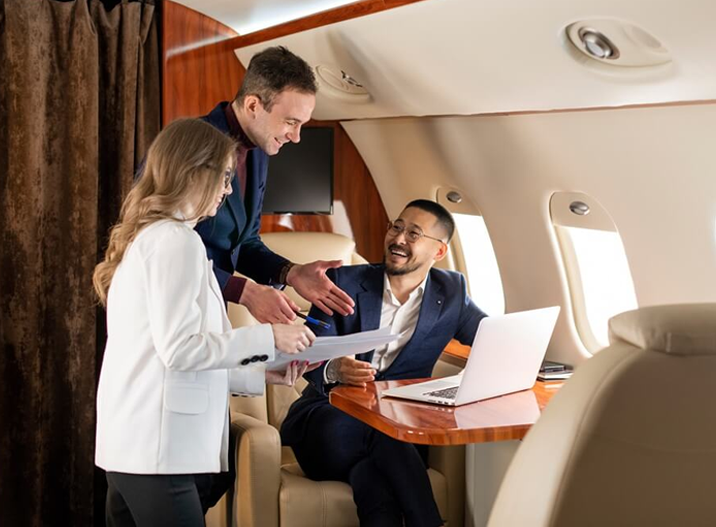 Three business professionals engaged in discussion inside a private jet with a laptop and documents on a wooden table.