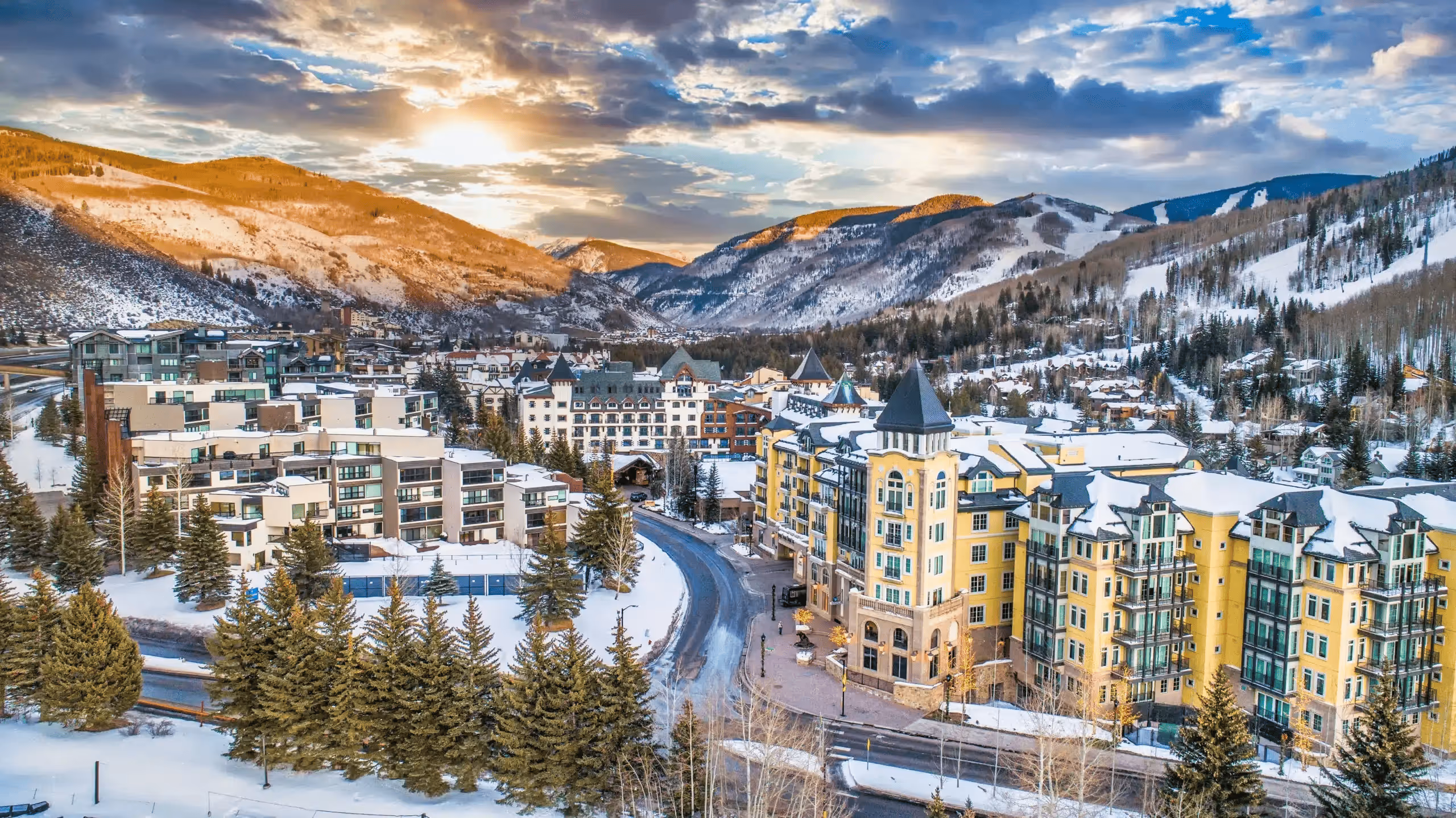 Snow-covered mountain town with buildings, pine trees, and a winding road under a cloudy sky at sunset.