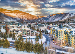 Snow-covered mountain town with buildings, pine trees, and a winding road under a cloudy sky at sunset.