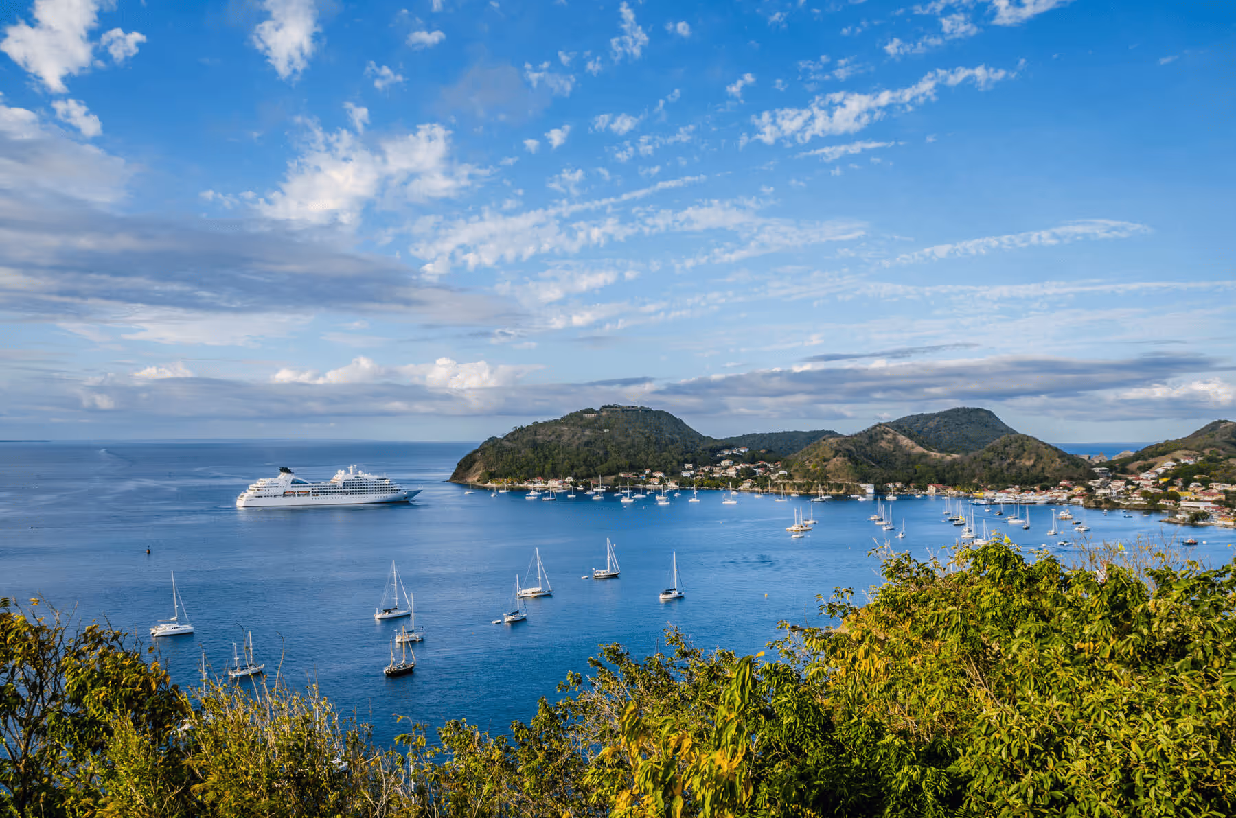 Scenic bay with several sailboats and a large cruise ship near a green, hilly coastline under a partly cloudy sky.