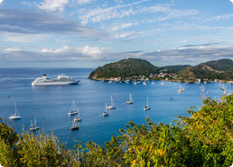 Scenic bay with several sailboats and a large cruise ship near a green, hilly coastline under a partly cloudy sky.