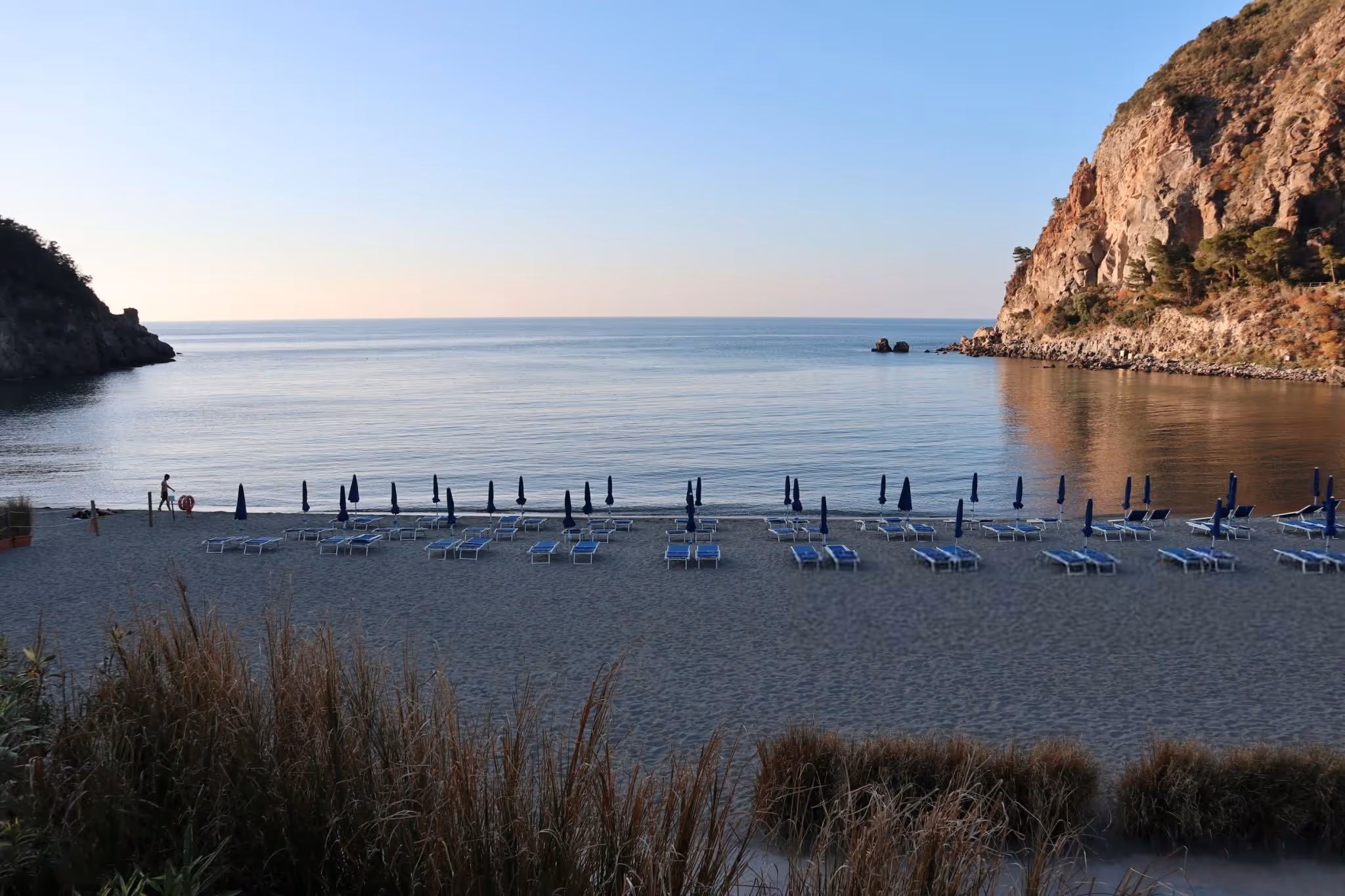Empty beach with rows of blue lounge chairs facing calm sea and rocky cliffs under a clear sky.