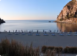 Empty beach with rows of blue lounge chairs facing calm sea and rocky cliffs under a clear sky.