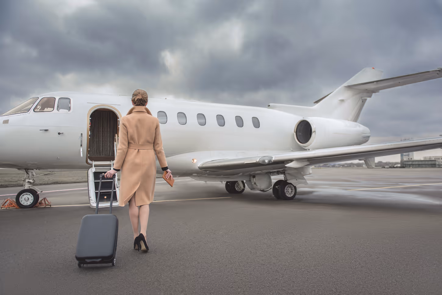 Woman in a tan coat with a rolling suitcase walking toward an open door of a private jet on an airport tarmac under a cloudy sky.