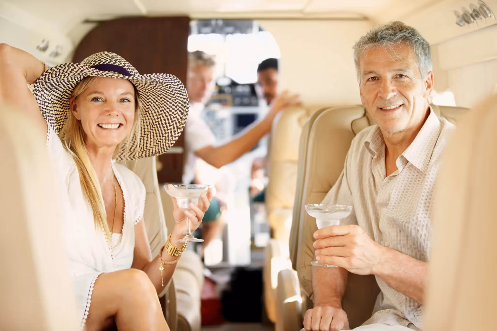 Smiling middle-aged man and woman sitting in an airplane cabin holding drinks, with other passengers in the background.