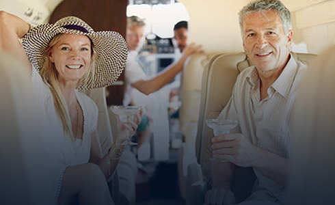 Smiling middle-aged man and woman sitting in an airplane cabin holding drinks, with other passengers in the background.