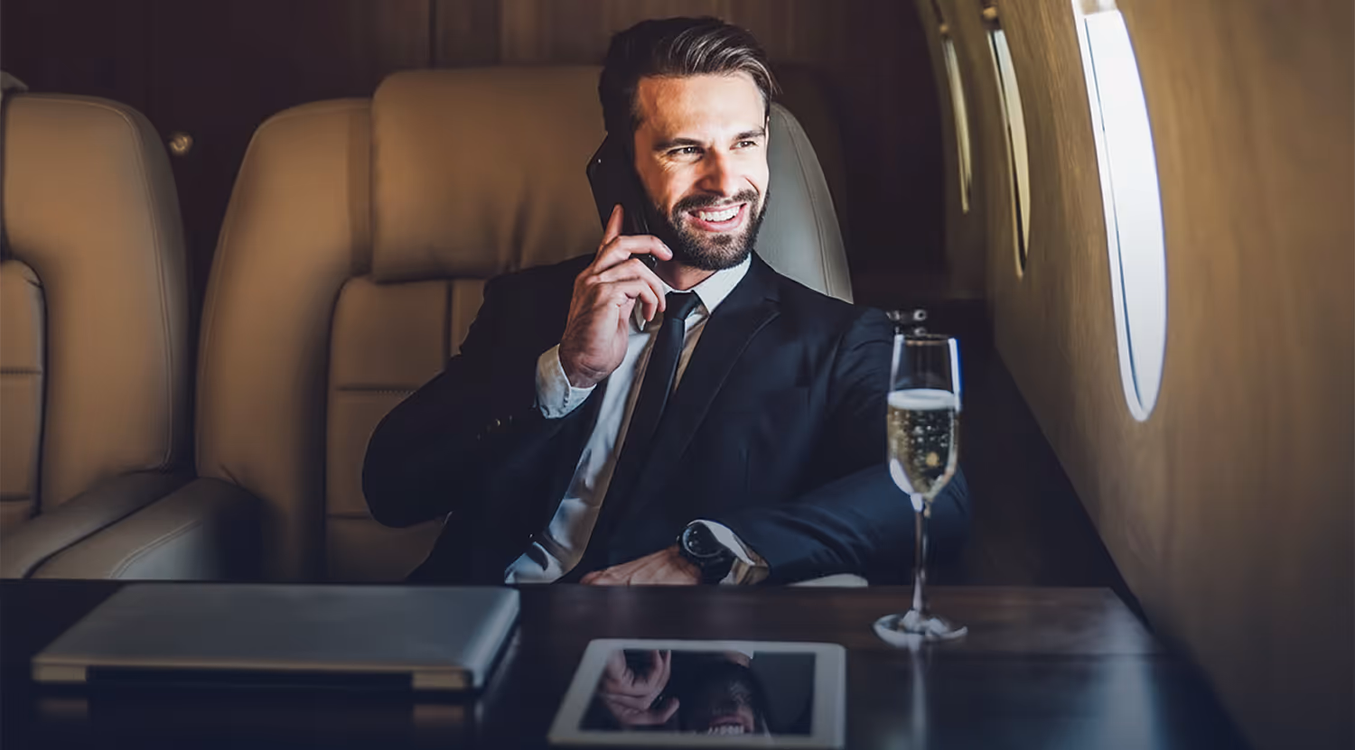 Smiling man in a suit talking on a phone inside a private jet, with a glass of champagne and electronic devices on the table.
