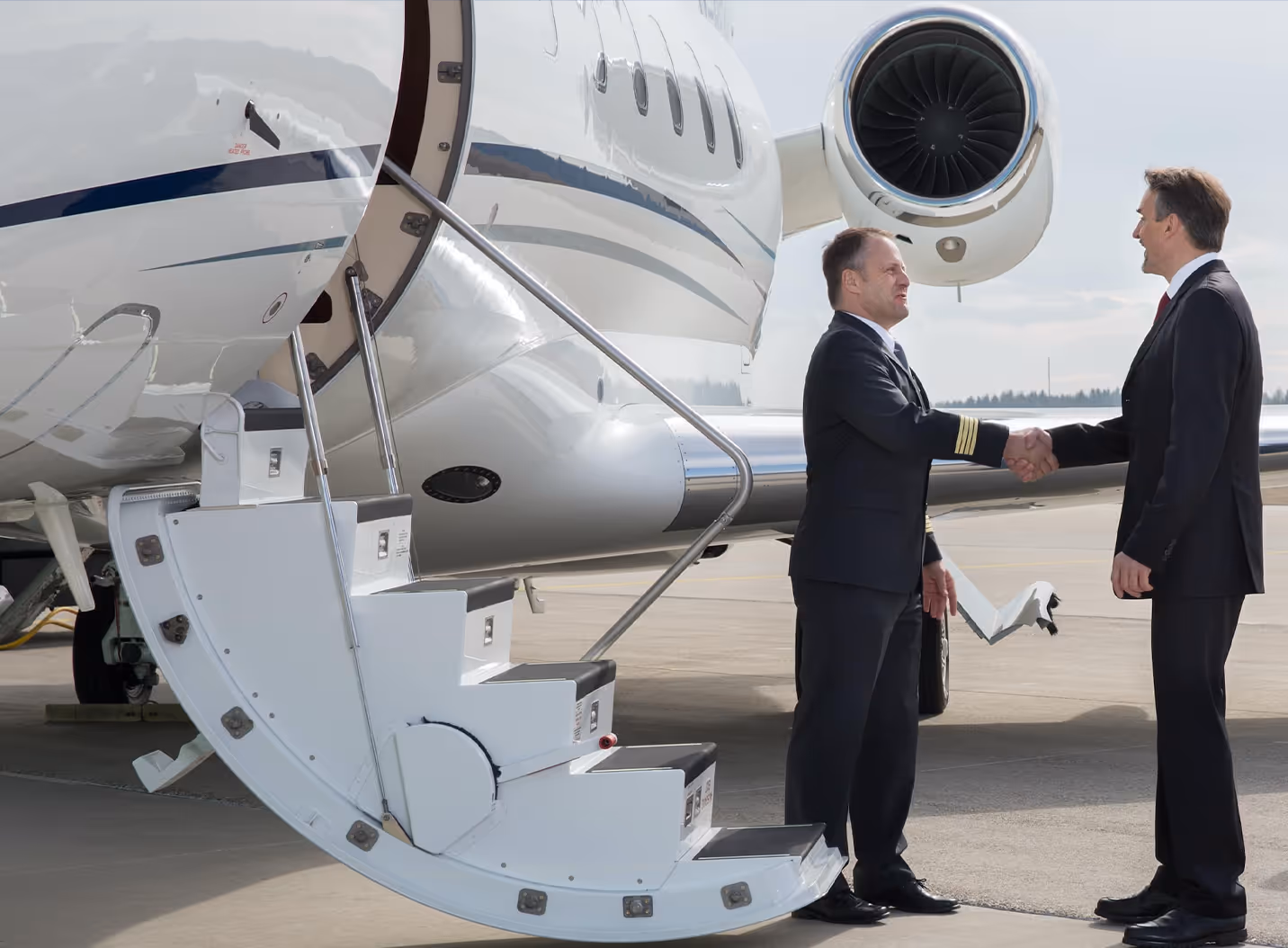 Two men, one in a pilot uniform and the other in a suit, shaking hands beside a private jet with its stairs extended on an airport tarmac.