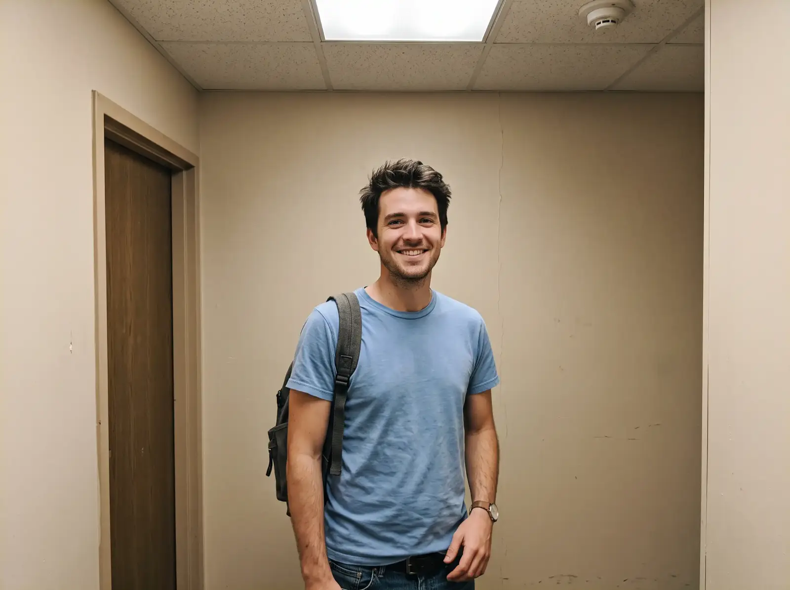 A man with a backpack standing against a plain beige hotel corridor wall