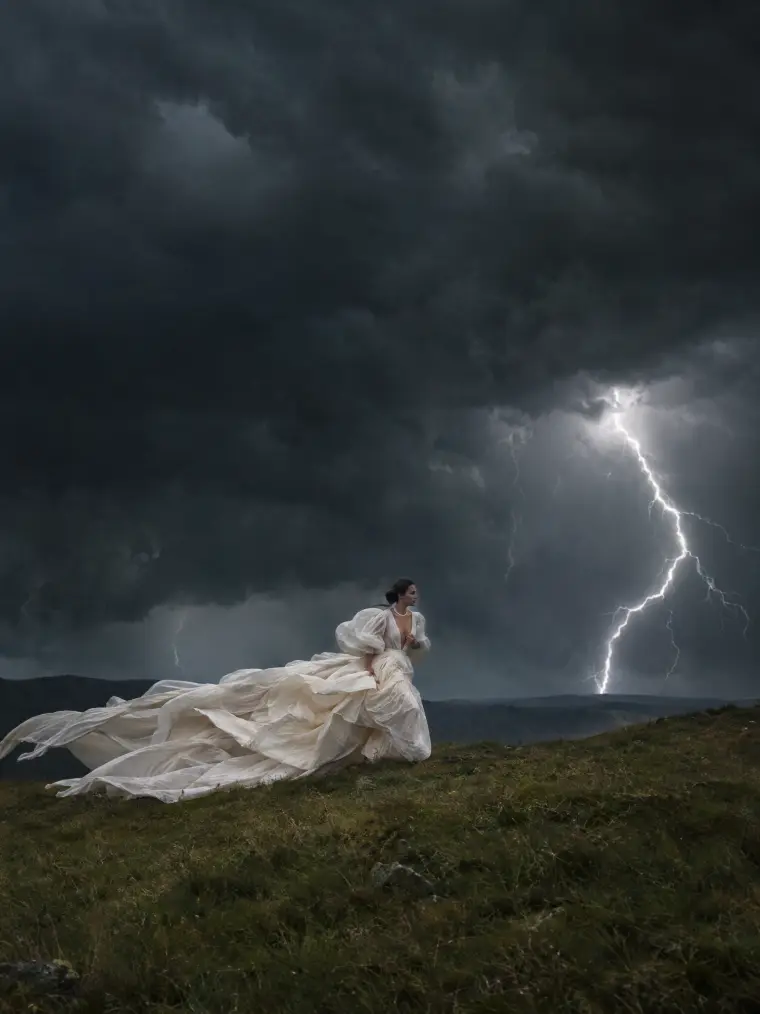 Woman in an ivory couture gown running across a hill during a thunderstorm