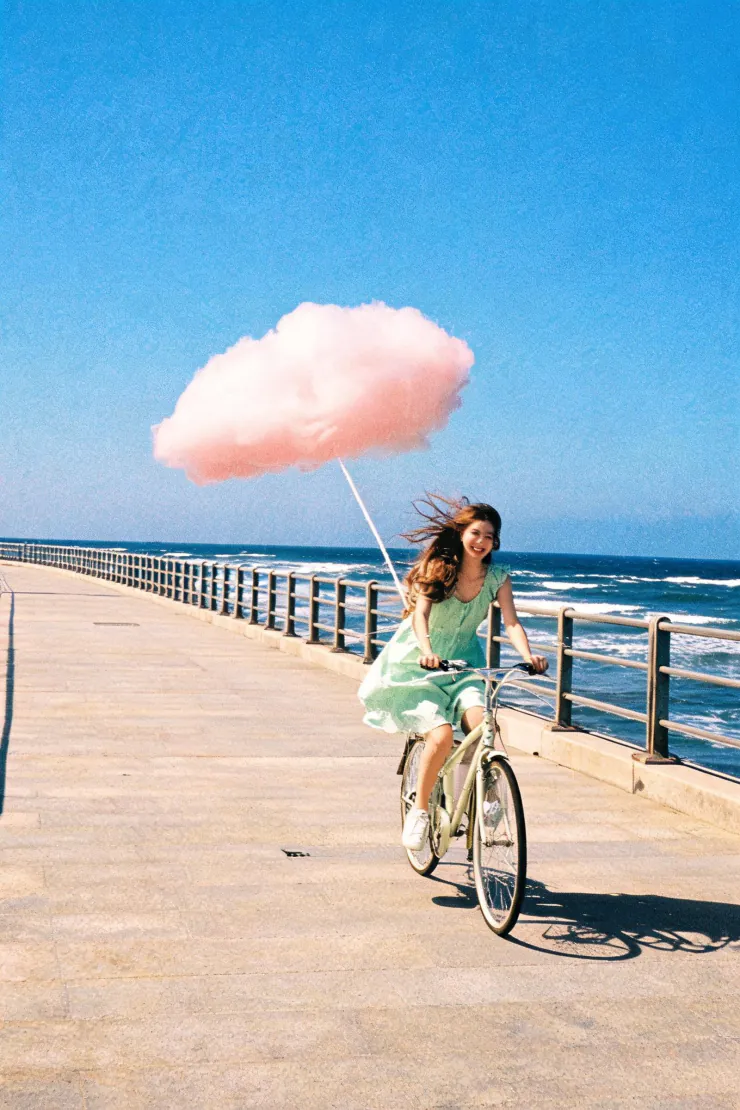 Girl on a bicycle towing a pink cloud along a seaside promenade