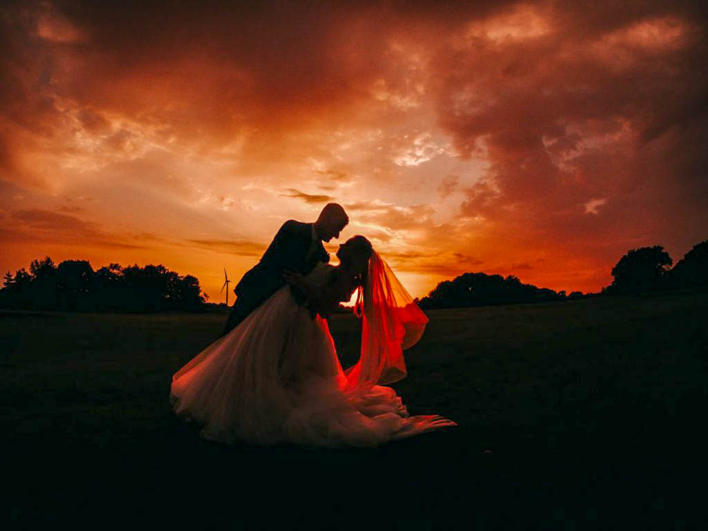 A couple silhouetted at sunset, bride's veil glowing red as groom dips her in a romantic embrace.