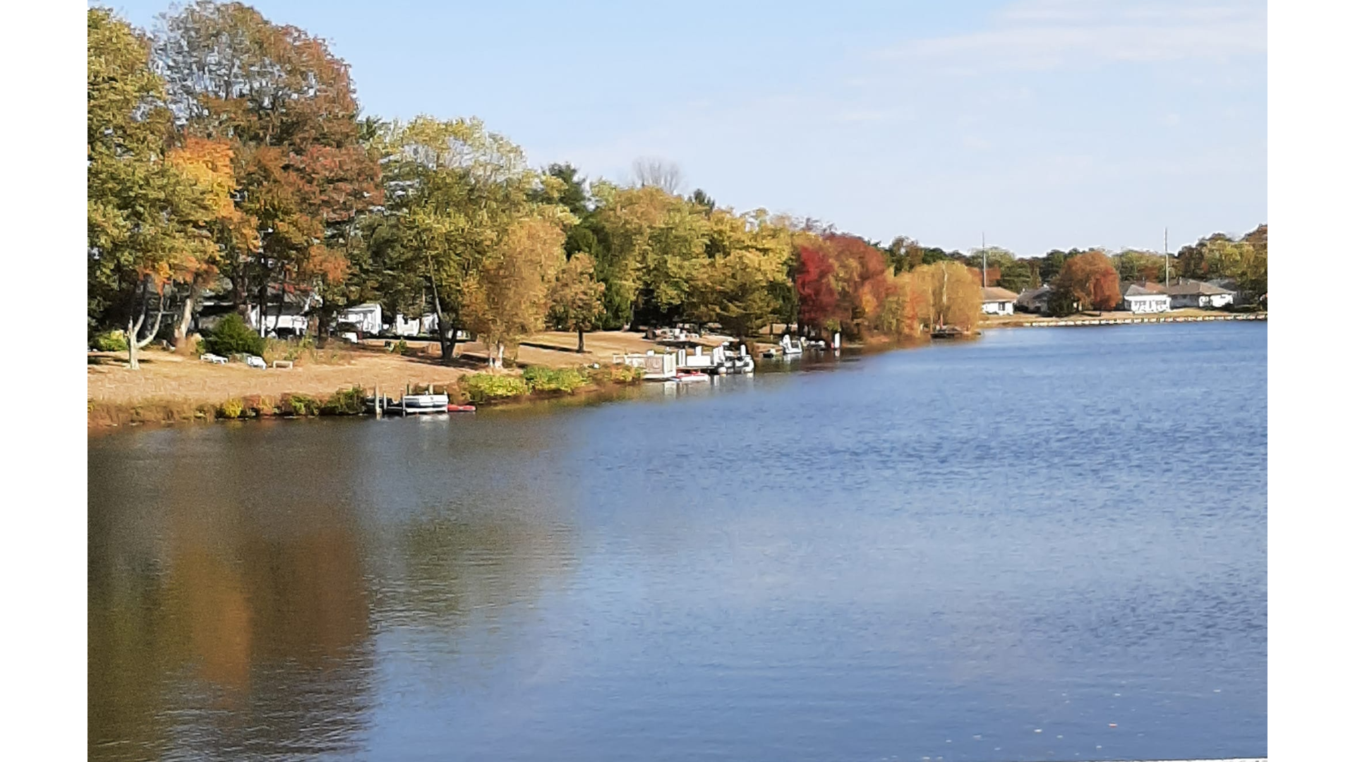 Scenic view of the waterfront community in Holiday City, Silverton, NJ, featuring a calm lake lined with small docks, pontoon boats, and vibrant autumn foliage among residential homes.
