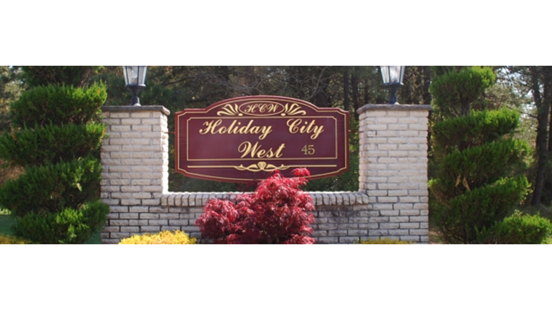 Brick entrance sign for Holiday City West, a 55+ adult community in Toms River, NJ, featuring a maroon and gold sign with decorative lighting, surrounded by manicured shrubs and colorful landscaping.