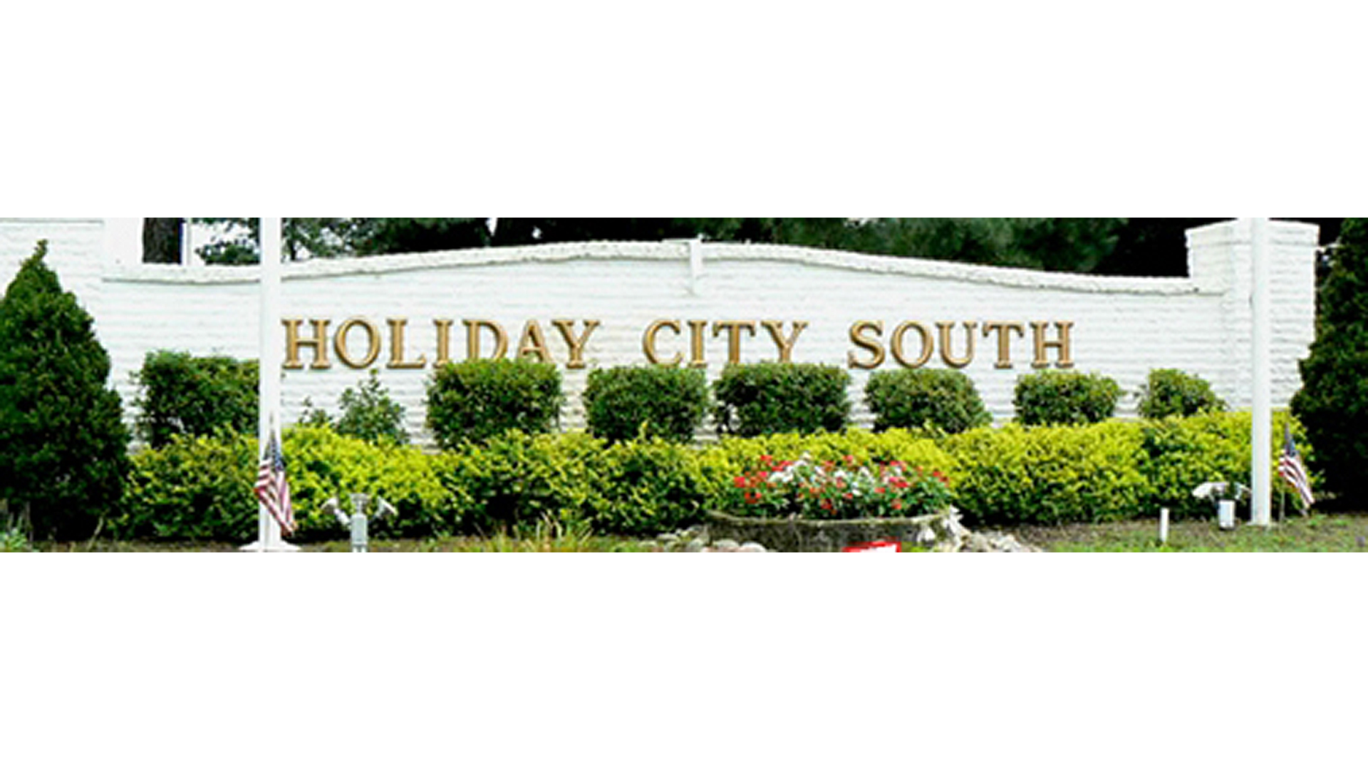 Landscaped brick entrance sign for Holiday City South, a 55+ adult community in Toms River, NJ, featuring gold lettering on a white wall, surrounded by manicured bushes, American flags, and flowerbeds.