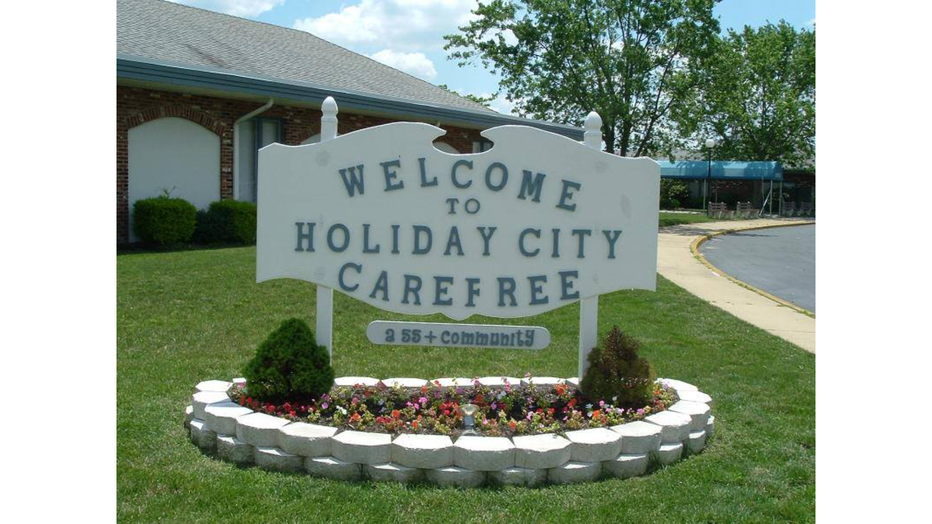 Welcome sign for Holiday City Carefree, a 55+ adult community in Toms River, NJ, featuring white carved lettering on a decorative sign surrounded by flower beds, green lawn, and a brick clubhouse in the background.