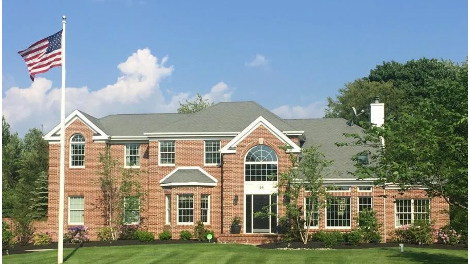 Brick colonial-style home in Manalapan, NJ with a gray shingle roof, lush lawn, and American flag. J.A.Y. Construction is a top choice for roofing and siding services in Monmouth County.