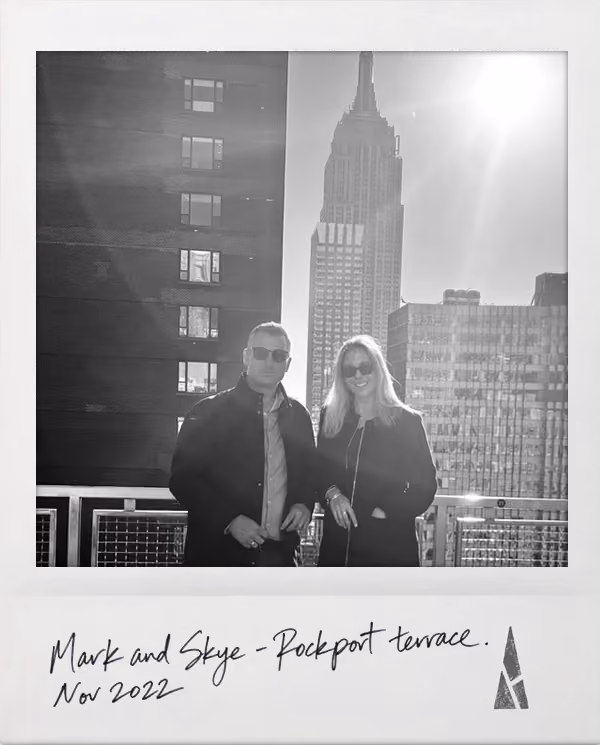 Polaroid picture of Mark and Skye wearing sunglasses holding a cake outdoors with mountains in the background