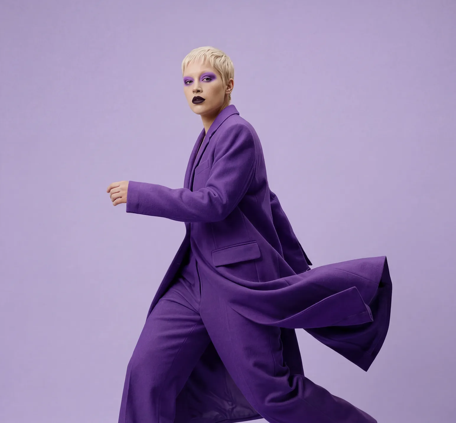 Person with short blonde hair wearing a flowing purple coat and matching pants walking against a purple background.