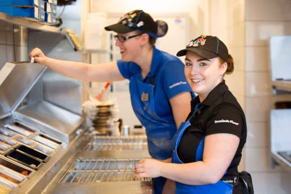 Two Domino's employees in the kitchen, smiling at the camera.