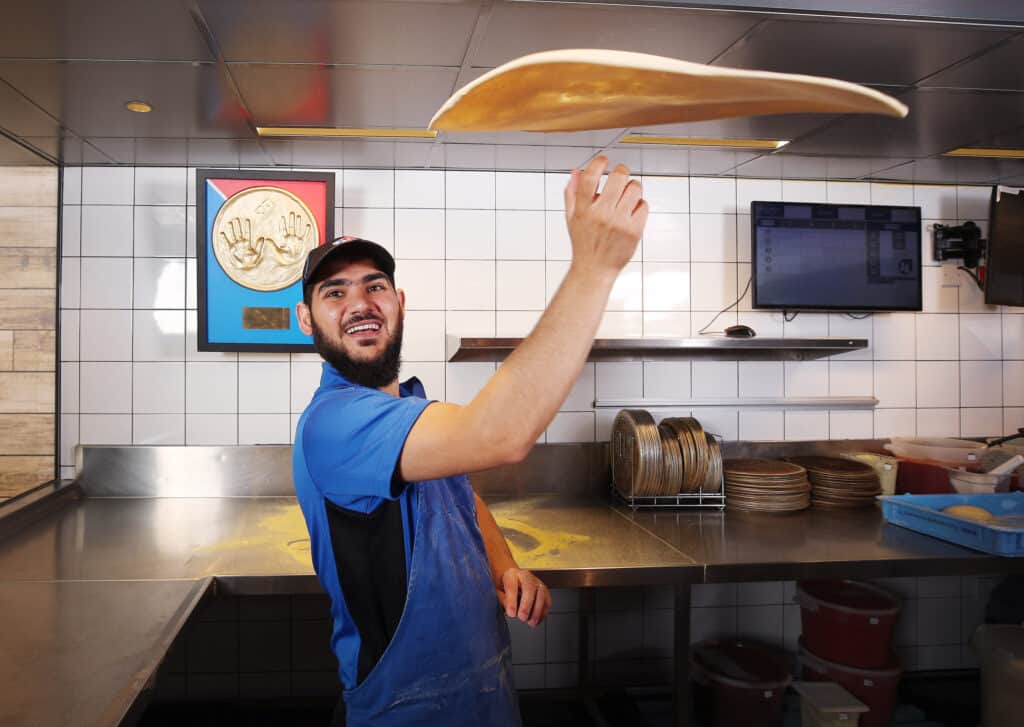 Domino's chef in kitchen spinning pizza base in air, while smiling at the camera.