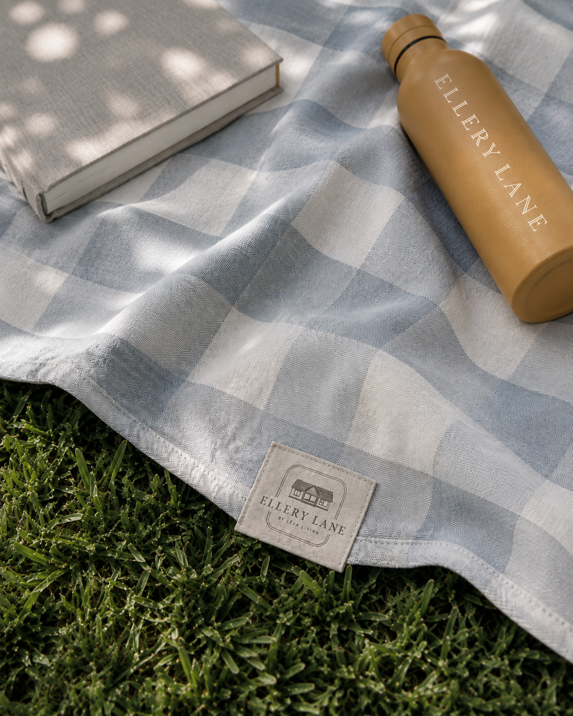 Ellery Lane branded picnic blanket in a soft blue and white check, styled on green grass with a mustard-toned water bottle and neutral linen-bound book.