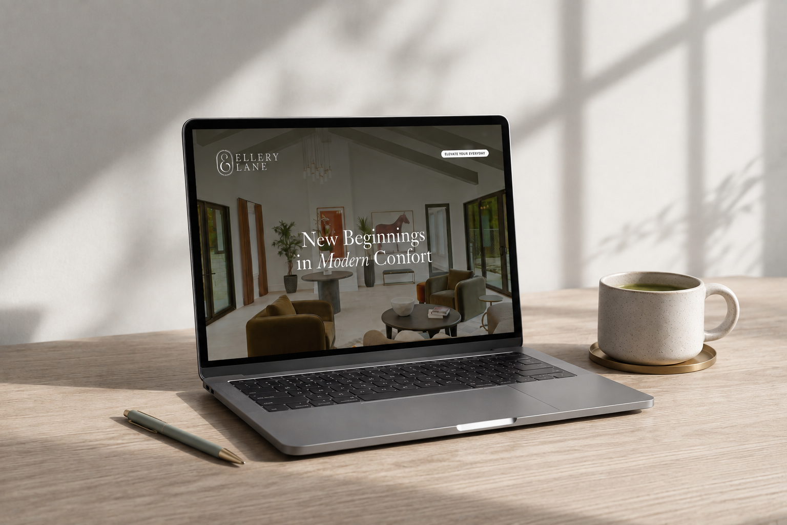 Ellery Lane website displayed on a laptop atop a pale white oak table, styled with a sage green pen and ceramic mug on a brass coaster in soft morning light.
