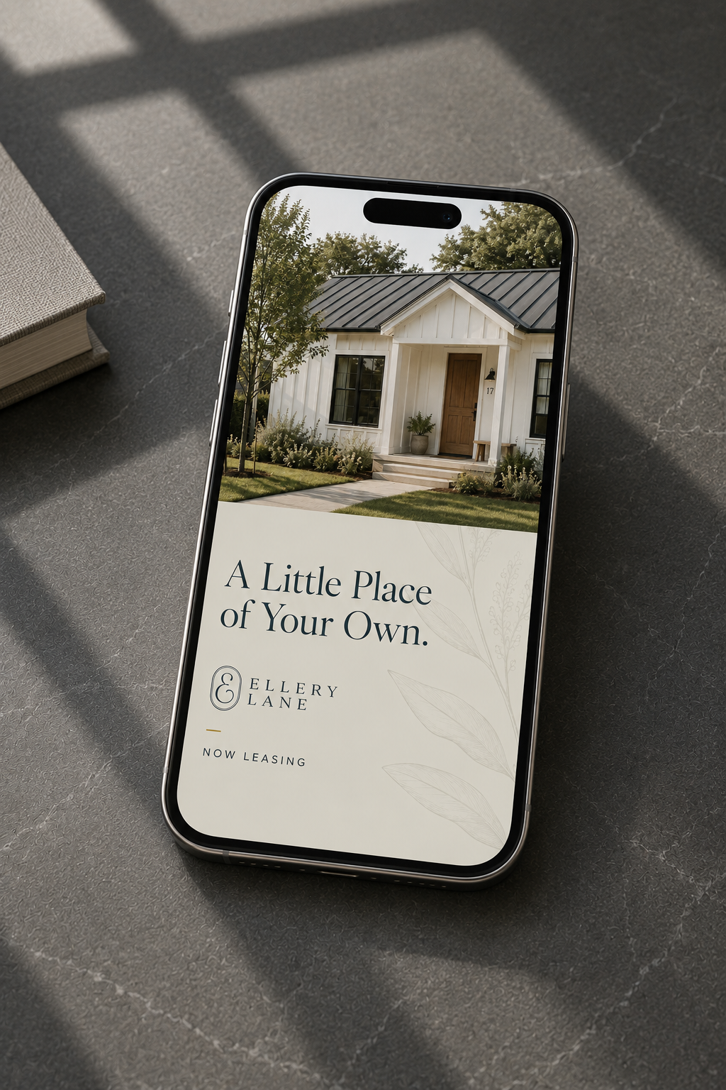 Ellery Lane social media advertisement displayed on an iPhone resting on a dark gray stone surface, featuring a white cottage and the message “A Little Place of Your Own.”