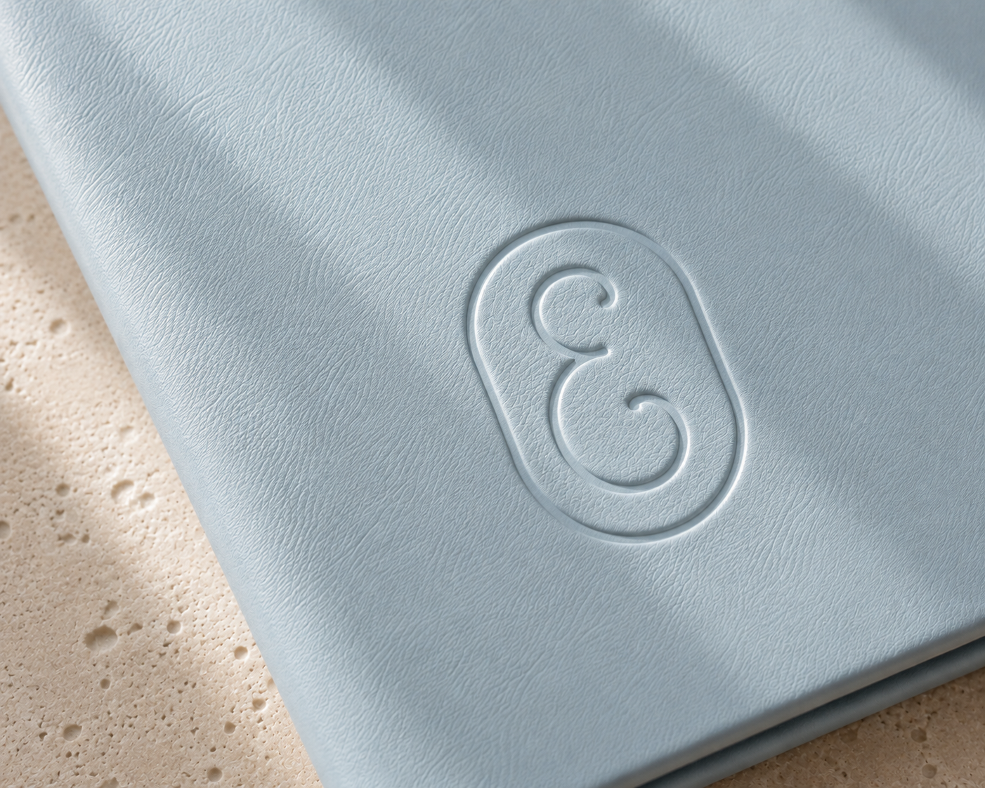 Close-up of a pale blue Ellery Lane notebook with the monogram blind embossed into the textured cover, resting on a light natural stone surface.
