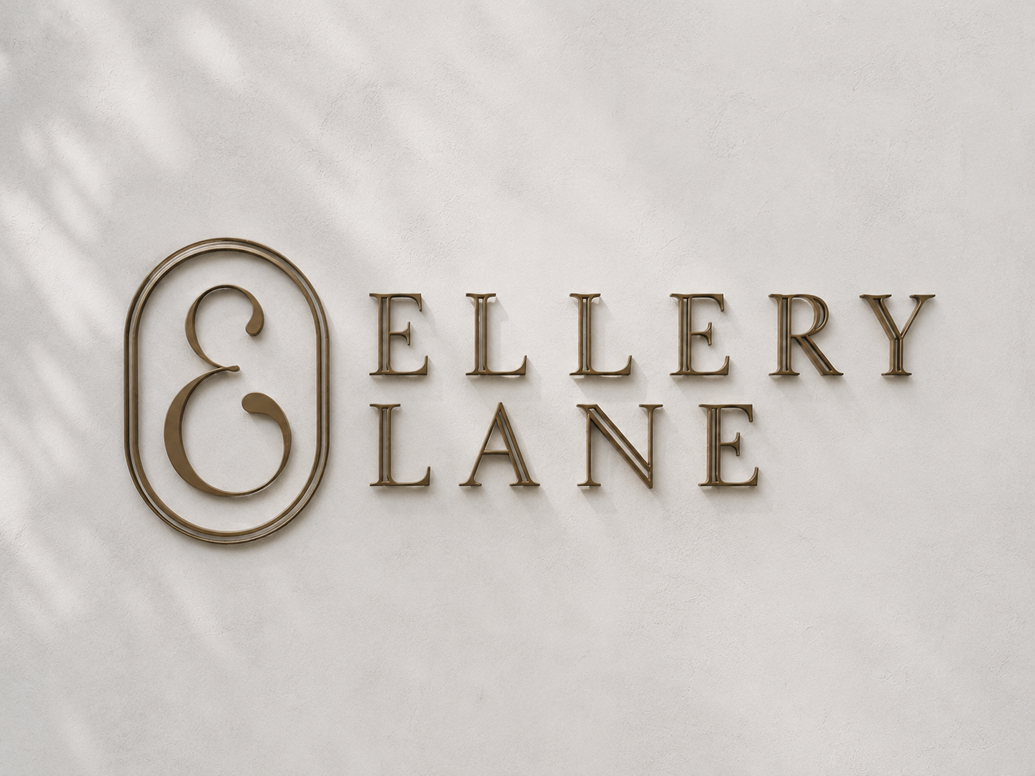 Bronze Ellery Lane logo mounted on a softly textured white plaster wall, illuminated by subtle natural morning light and soft shadows.
