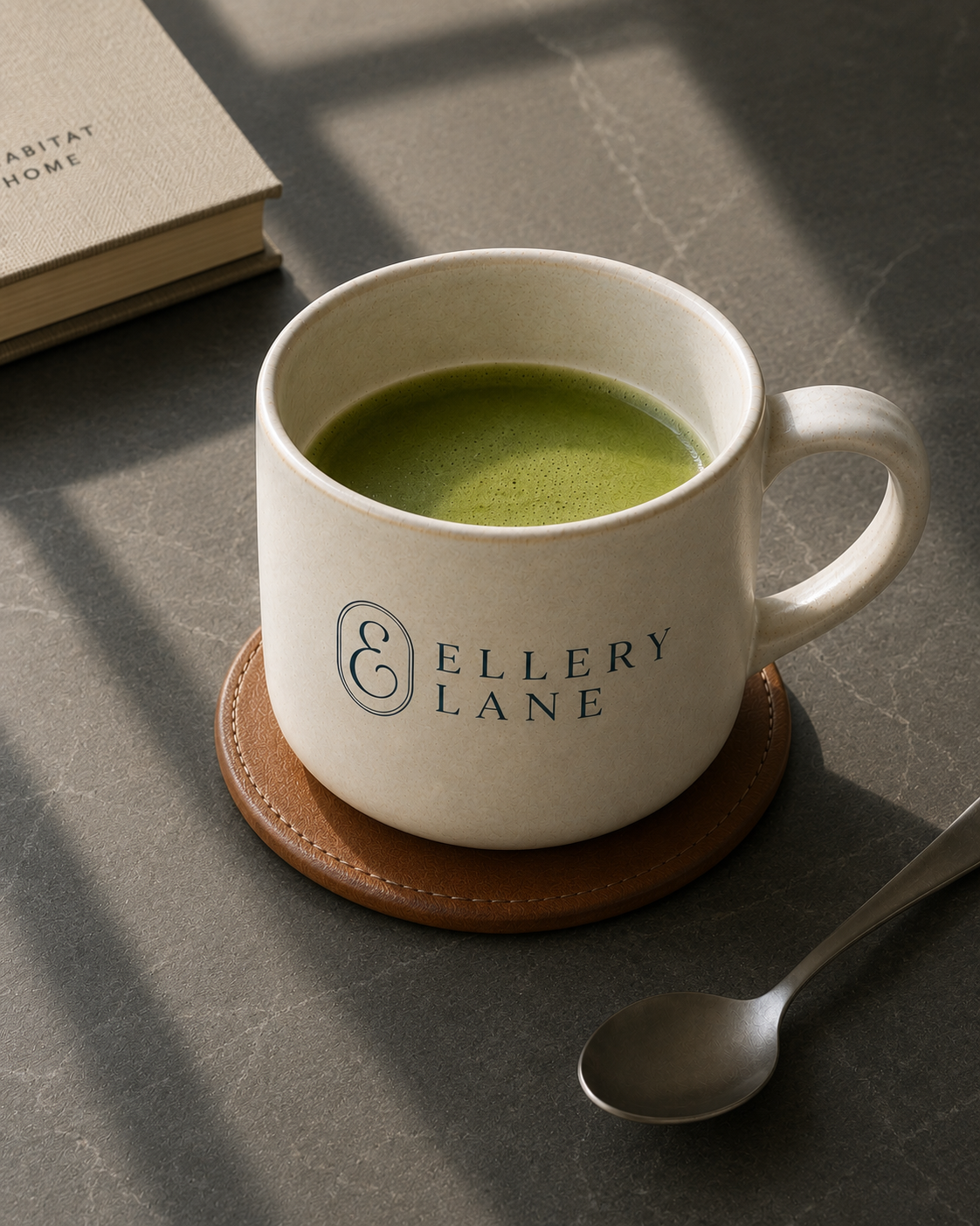 Ellery Lane ceramic mug filled with matcha and featuring the brand logo, set on a warm leather coaster atop a dark matte stone surface with a spoon and book nearby.