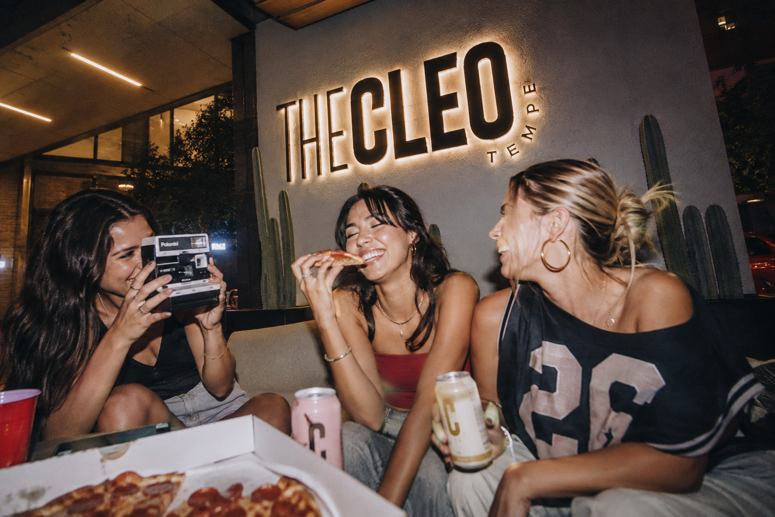 Three friends sharing pizza and drinks beneath an illuminated The Cleo Tempe logo in a lively apartment lounge.