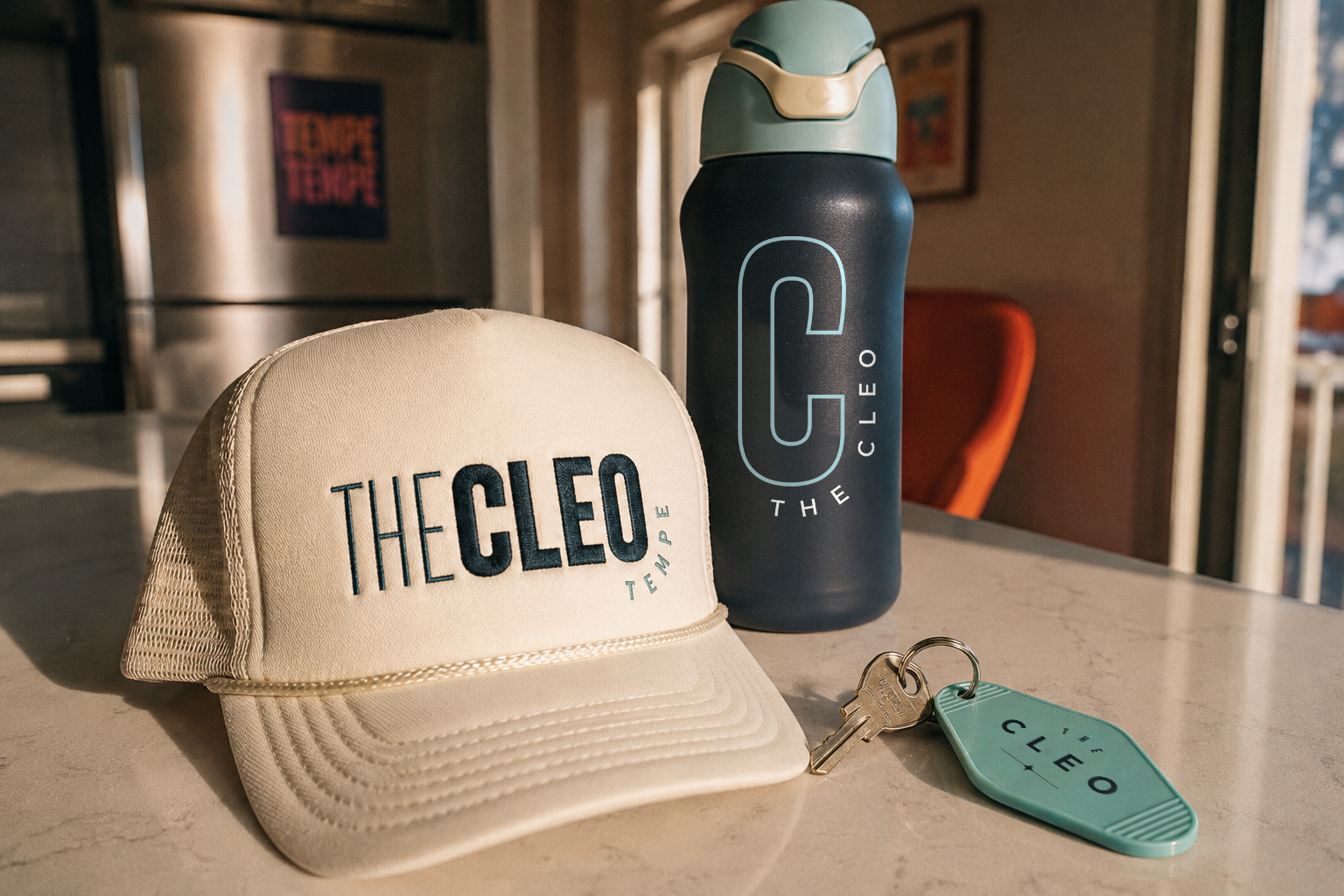 The Cleo Tempe branded merchandise, including a cream trucker hat, navy water bottle, and teal apartment key tag displayed in a sunlit interior.