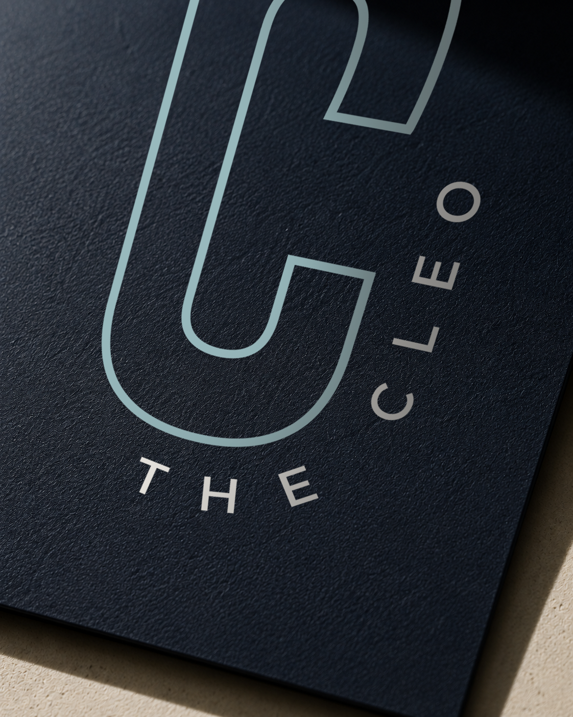 Close-up of The Cleo logo printed in pale blue and cream on textured navy material, highlighting the brand mark and typography.