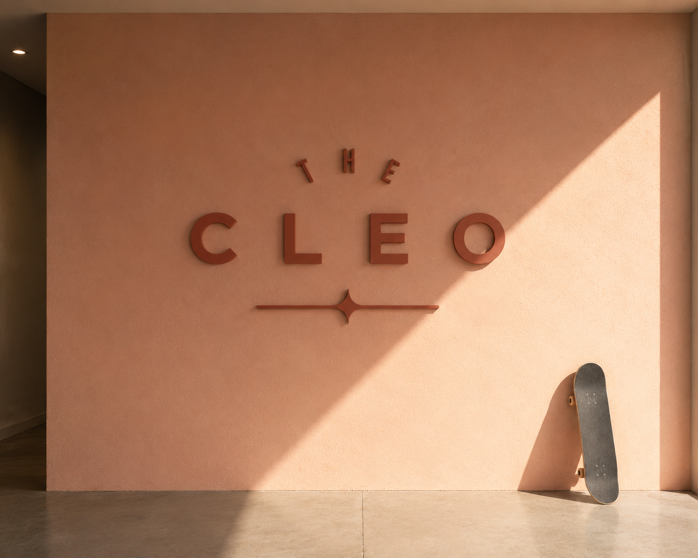 The Cleo dimensional logo displayed on a warm blush interior wall, with a skateboard and dramatic afternoon sunlight.
