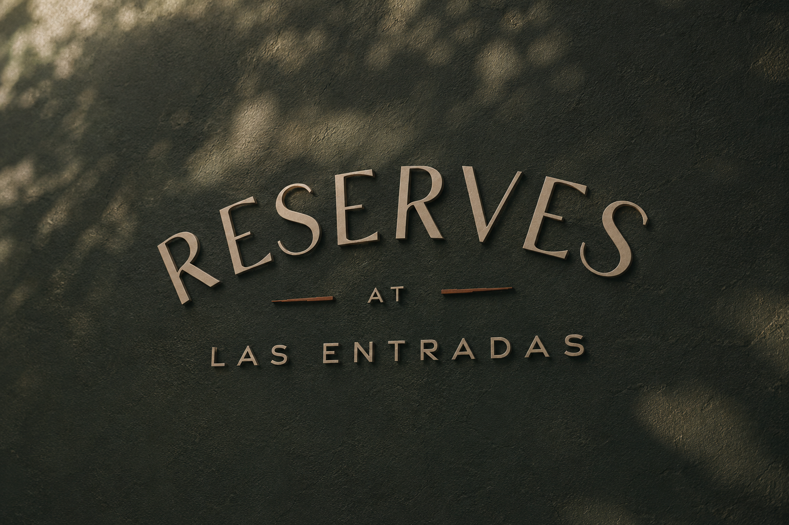 Reserves at Las Entradas dimensional wordmark mounted on a textured dark green wall, with soft tree shadows cast across the surface.
