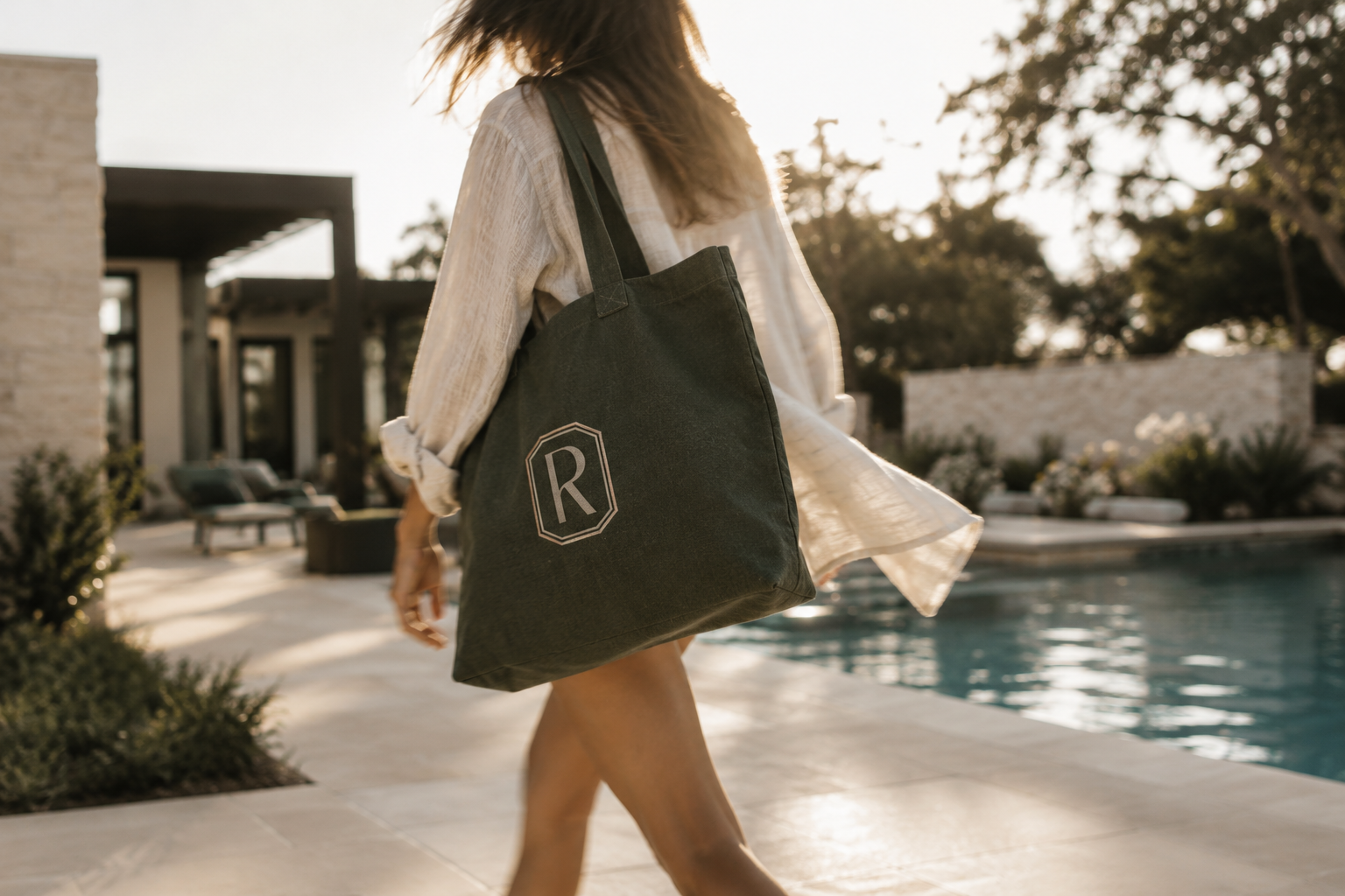 Woman carrying a Reserves at Las Entradas branded canvas tote while walking beside a luxury residential pool in warm Texas sunlight.