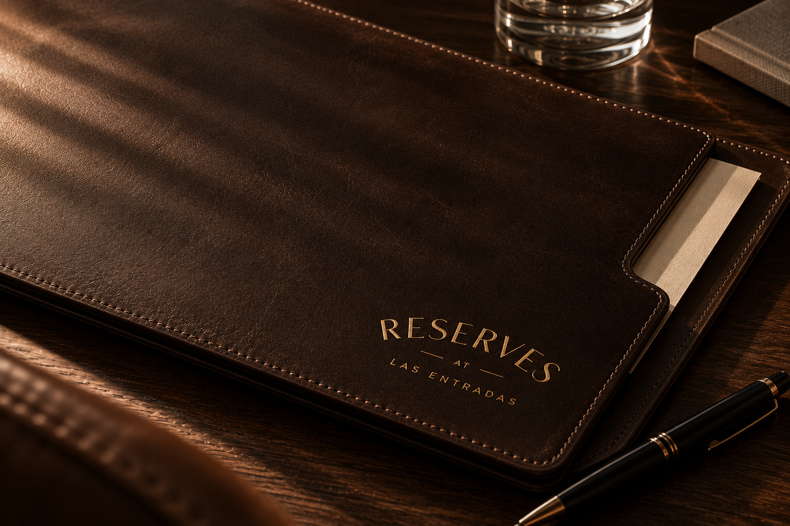 Dark brown leather Reserves at Las Entradas folio with gold foil branding and contrast stitching, styled with a black pen on a warm wood desk.