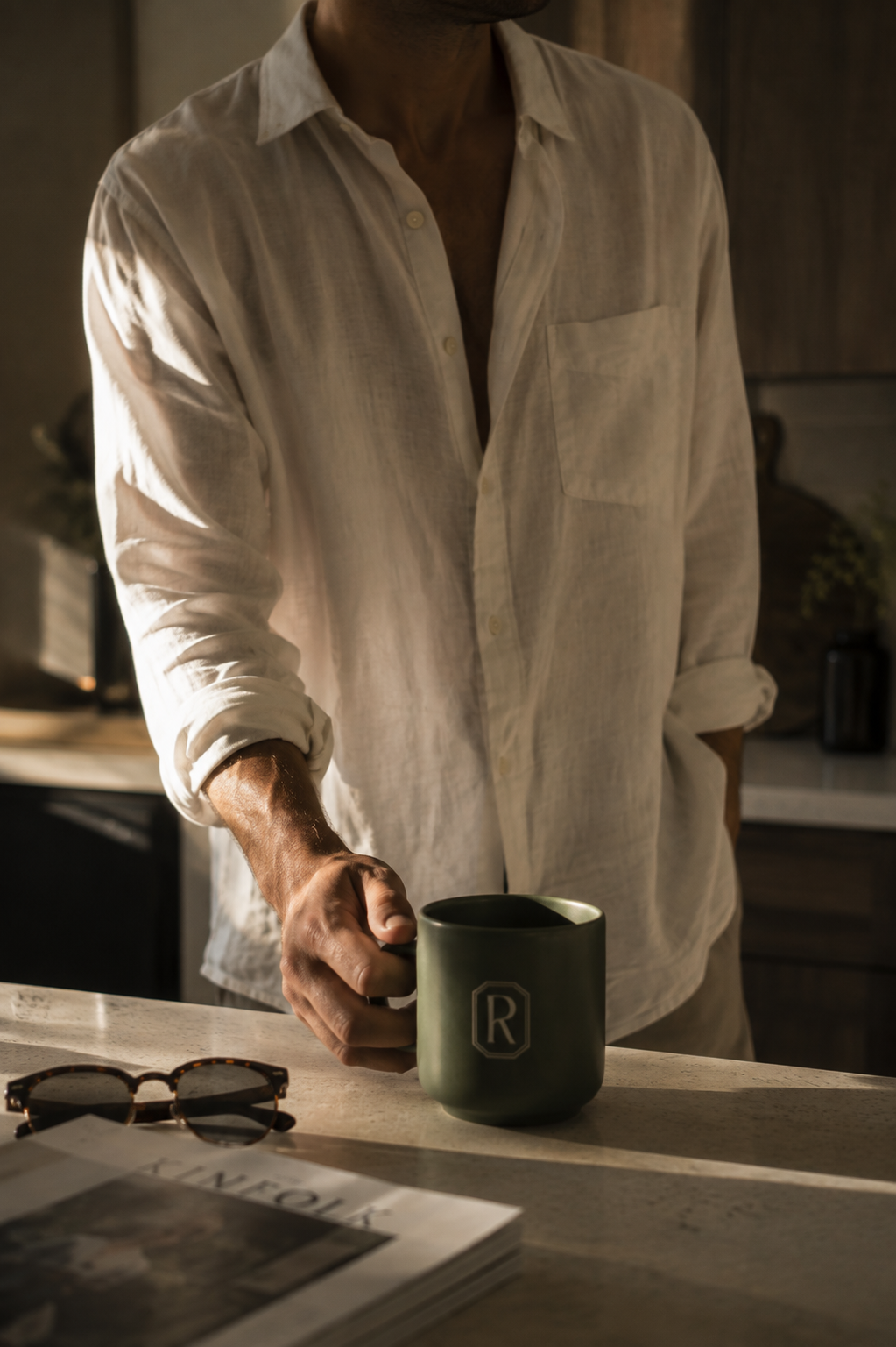 Man in a white linen shirt holding a dark green Reserves monogram coffee mug in a warmly lit modern kitchen.