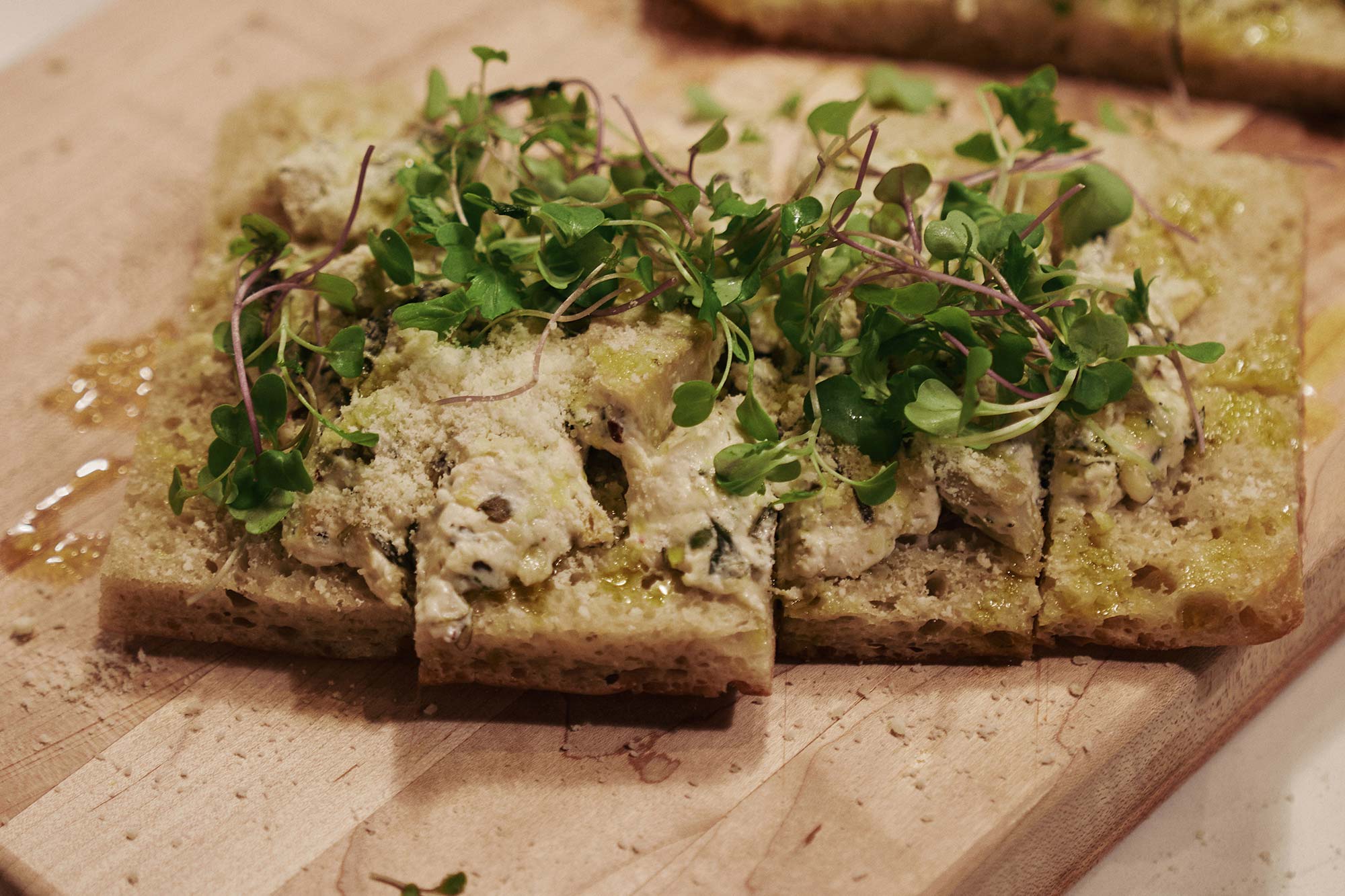 Close-up of sliced focaccia bread topped with creamy chicken, microgreens, and grated cheese on a wooden board.