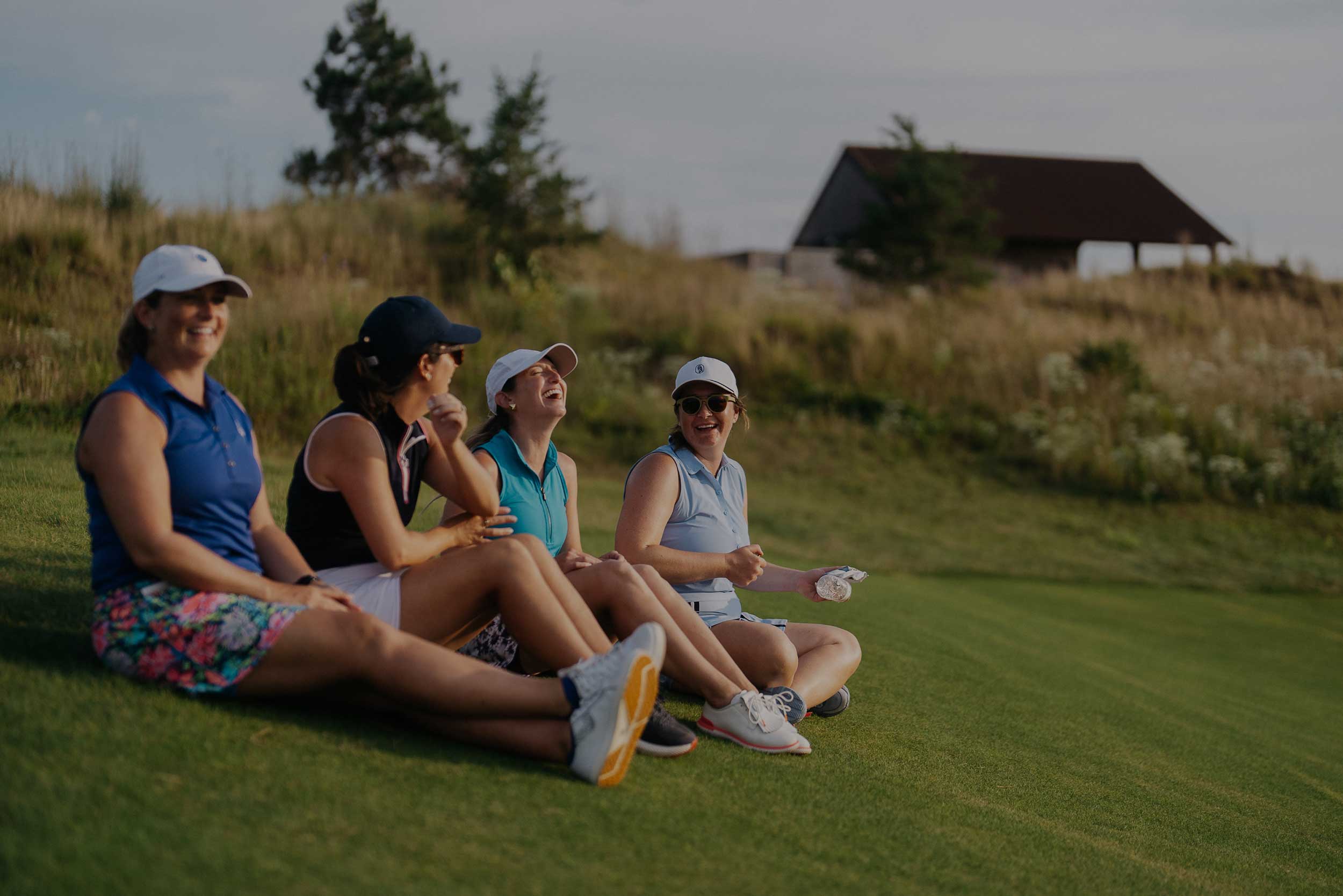 Four women golfers sitting on the grass, laughing and relaxing together after a round, with a scenic golf course and pavilion in the background.
