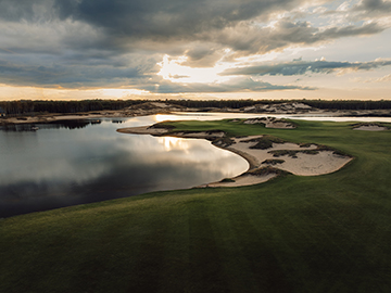 A serene golf course at sunset with sand traps, green fairways, and a calm water hazard reflecting the cloudy sky.