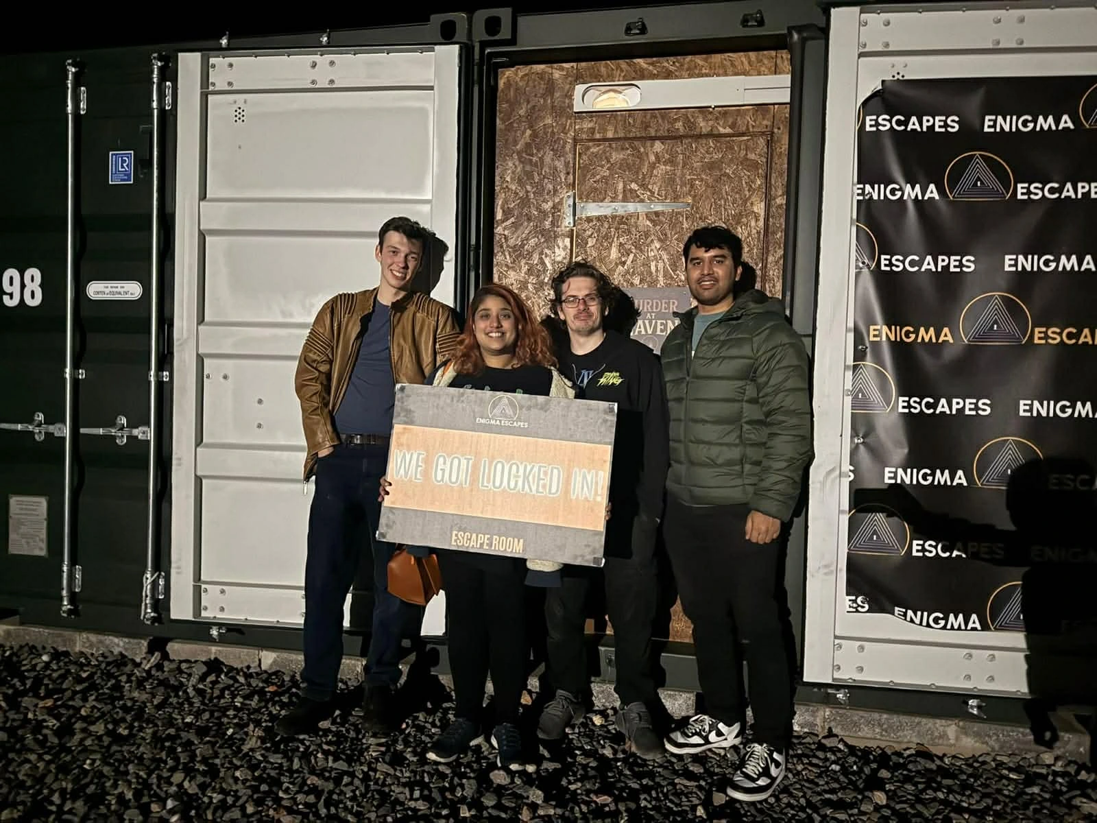 Group of four people standing outside an escape room container holding a sign that says 'We Got Locked In!'