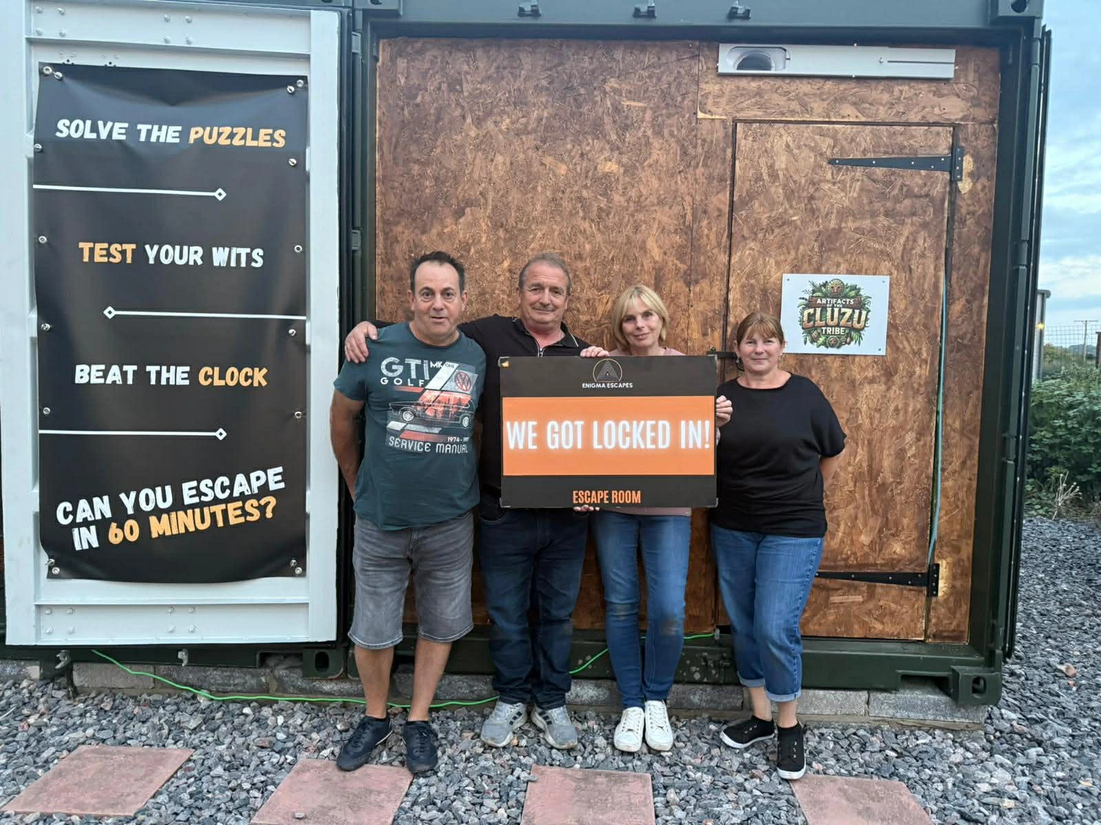 Four adults standing in front of a wooden door, holding a sign that reads 'We got locked in! Escape Room,' with a banner beside them promoting puzzles and a 60-minute escape challenge.